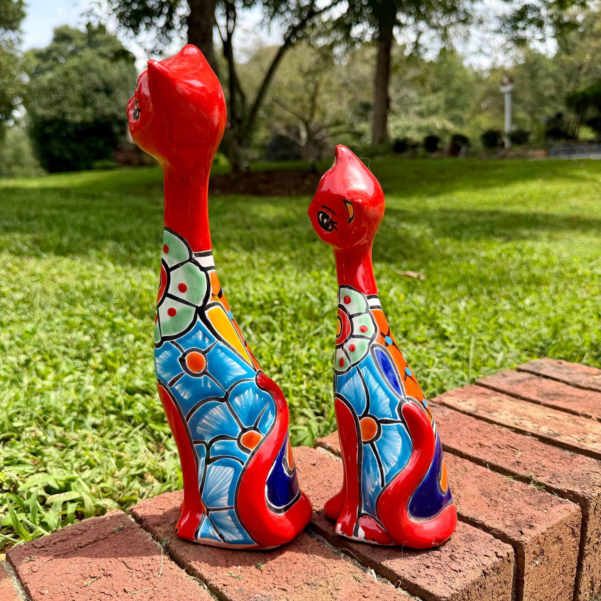 Two hand-painted ceramic cat figurines in vibrant red and multicolor patterns, displayed on a brick ledge in an outdoor garden setting. this is their right side view.