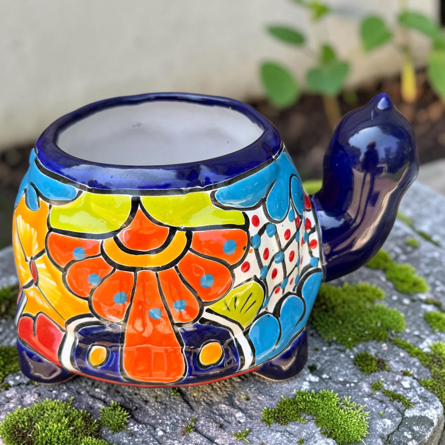 A colorful talavera turtle planter with a hand-painted Mexican-style pattern in orange, blue, yellow, and white, sitting on a mossy stone outdoors. Back of turtle planter.