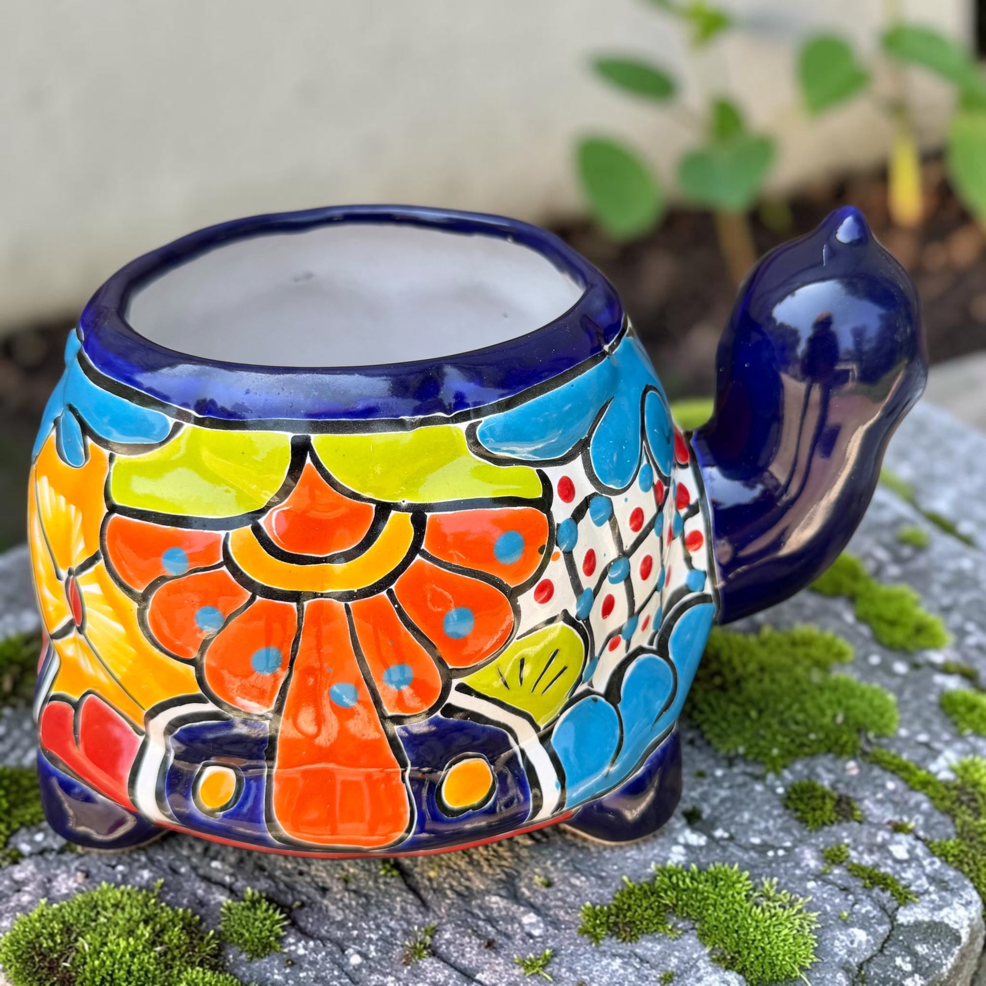 A colorful talavera turtle planter with a hand-painted Mexican-style pattern in orange, blue, yellow, and white, sitting on a mossy stone outdoors. Back of turtle planter.