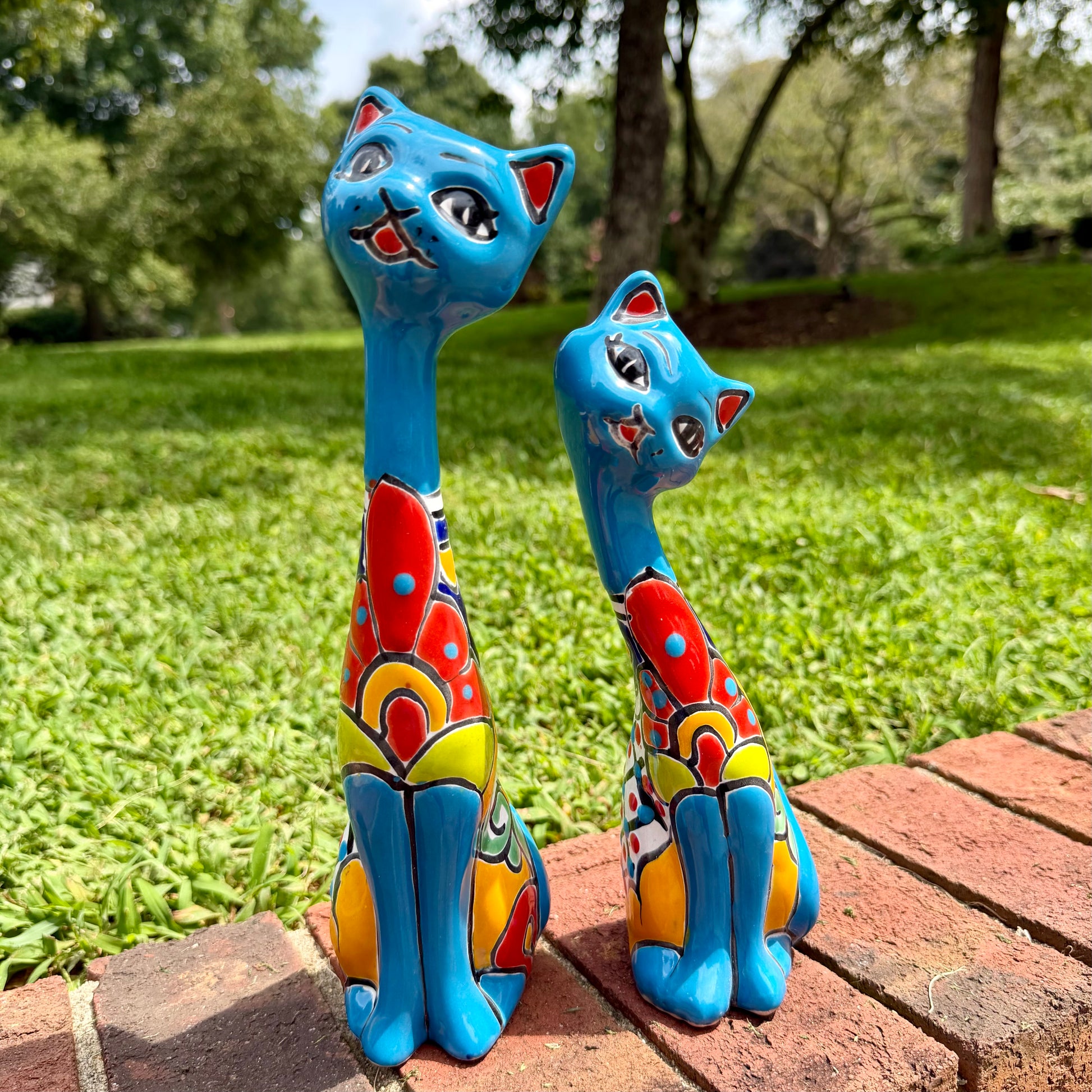 Two colorful turquoise hand-painted ceramic cat garden statues in blue, red, yellow, and white, displayed on a brick path in a grassy backyard.