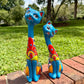 Two colorful turquoise hand-painted ceramic cat garden statues in blue, red, yellow, and white, displayed on a brick path in a grassy backyard.