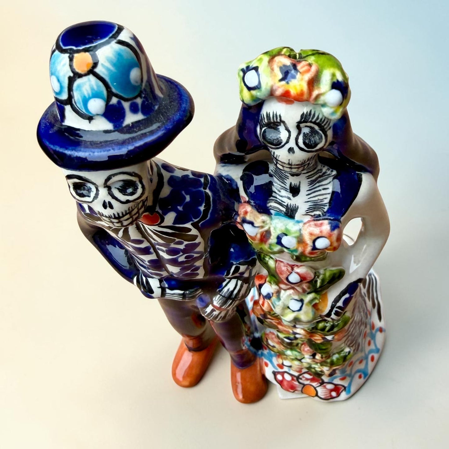 A hand-painted Talavera Day of the Dead bride and groom figurine. The groom is dressed in a blue suit with white patterns, a black bow tie, and an orange-red bow tie accent, wearing a blue hat. Top view looking down.