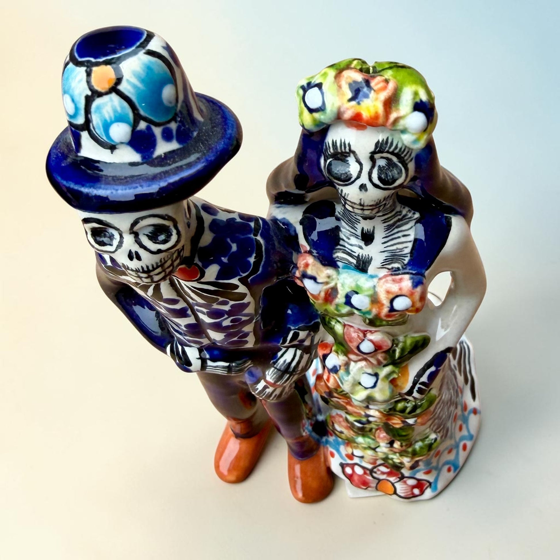 A hand-painted Talavera Day of the Dead bride and groom figurine. The groom is dressed in a blue suit with white patterns, a black bow tie, and an orange-red bow tie accent, wearing a blue hat. Top view looking down.