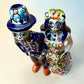 A hand-painted Talavera Day of the Dead bride and groom figurine. The groom is dressed in a blue suit with white patterns, a black bow tie, and an orange-red bow tie accent, wearing a blue hat. Top view looking down.