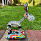 A colorful hand-painted Talavera Catrina donkey figurine, standing on a colorful floral base in an outdoor backyard setting, left side view of donkey.