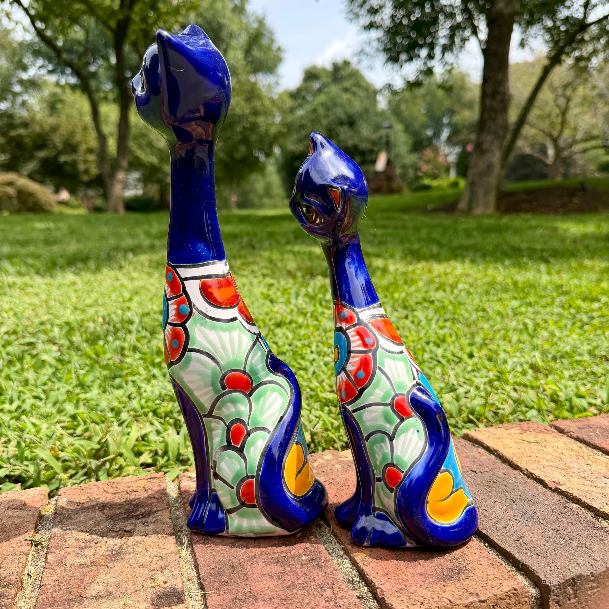 Two colorful ceramic cat garden statues in a lush green backyard setting, standing on a brick path.