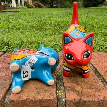 Two hand-painted Mexican-style ceramic cat figurines, one blue and one red, decorated with colorful geometric patterns, standing on a brick surface outdoors bottom view.