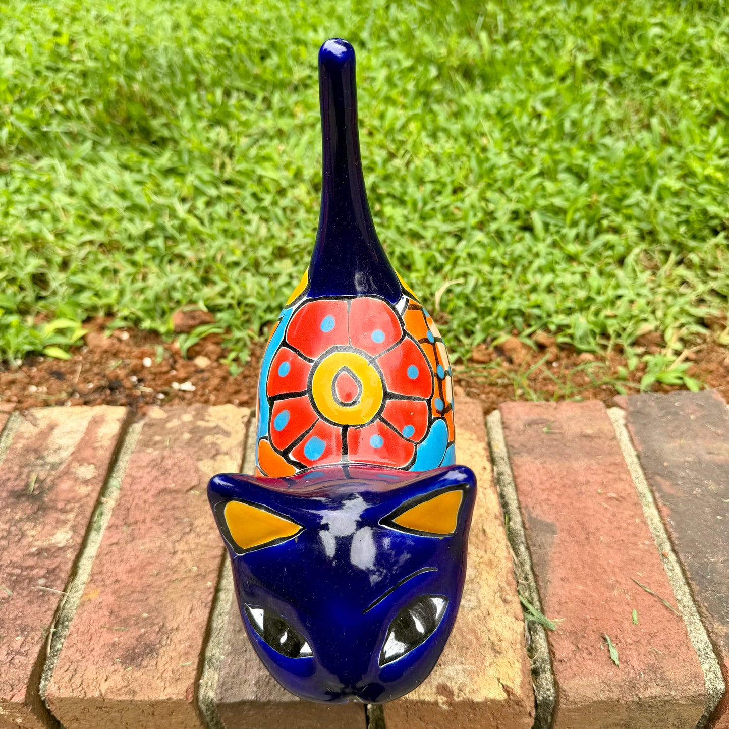 A glossy talavera ceramic cat figurine decorated with vibrant hand-painted patterns in blue, orange, yellow, and red, standing on a brick path with a green lawn and a house in the blurred background. View from the top.