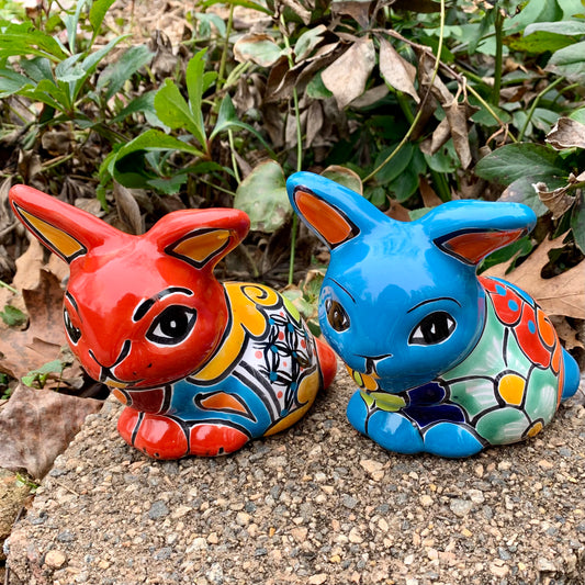Two colorful Mexican Talavera rabbit figurines on a stone surface with greenery in the background Red and Turquoise