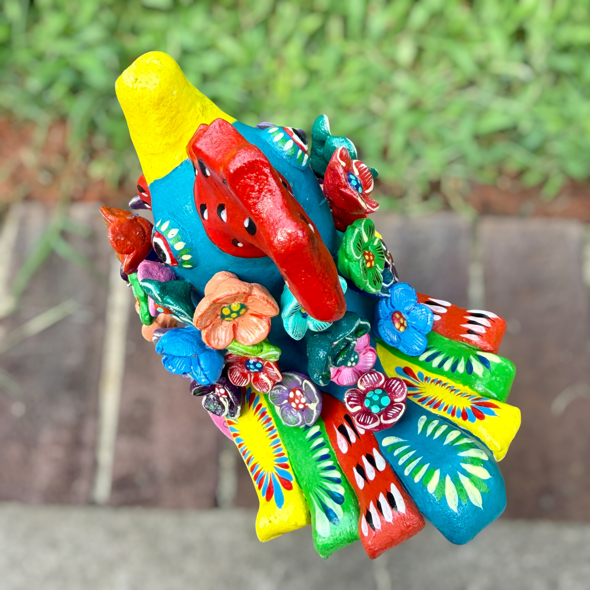 A colorful decorative rooster folk art sculpture with a blue body, yellow legs and beak, and many bright flower ornaments, standing on a pink circular base outdoors on a brick path with a blurred green lawn behind it. Top view of Rooster. 