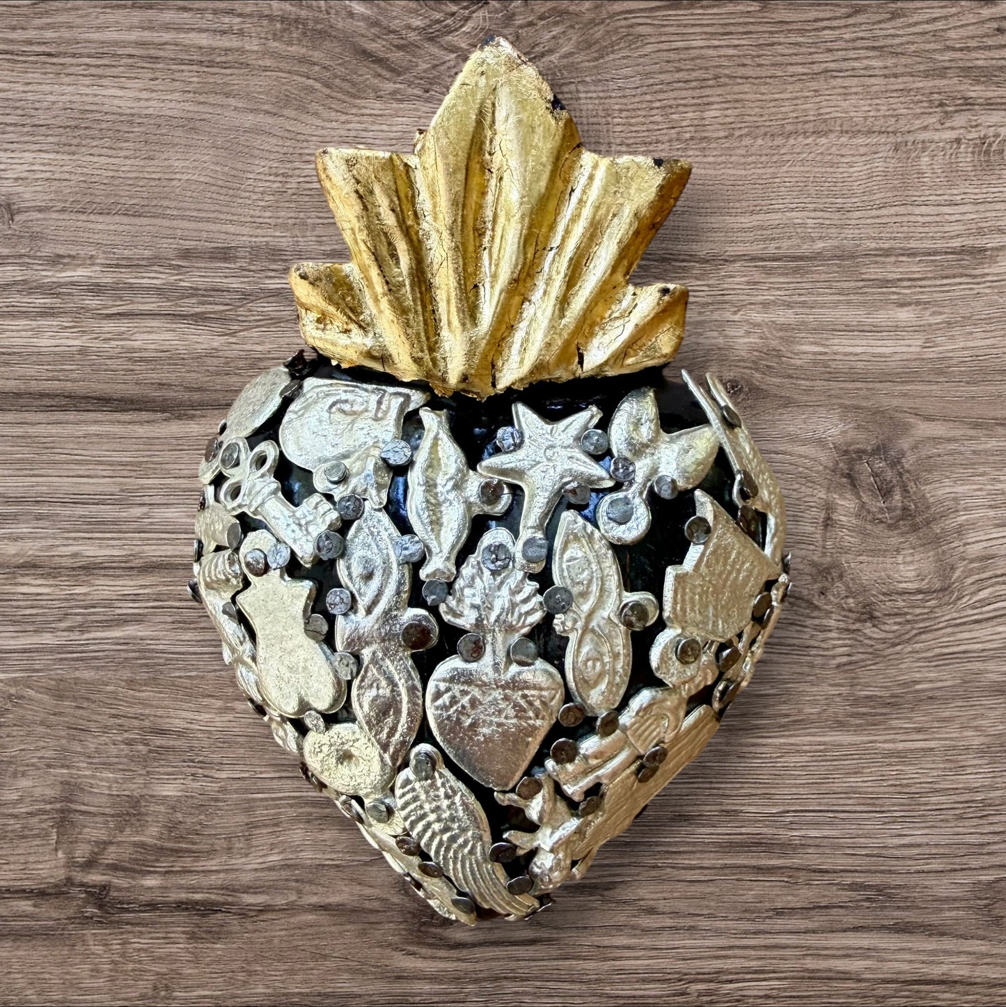A decorative Milagros Sacred Heart featuring a black and silver painted heart adorned with charms, set against a rustic wood background.