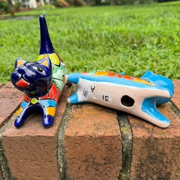 Two colorful hand-painted ceramic cats sitting on a brick ledge with a green lawn and trees in the background. One is cobalt  blue and the other is turquoise, both have traditional multi-color  talavera designs, bottom figurine view.