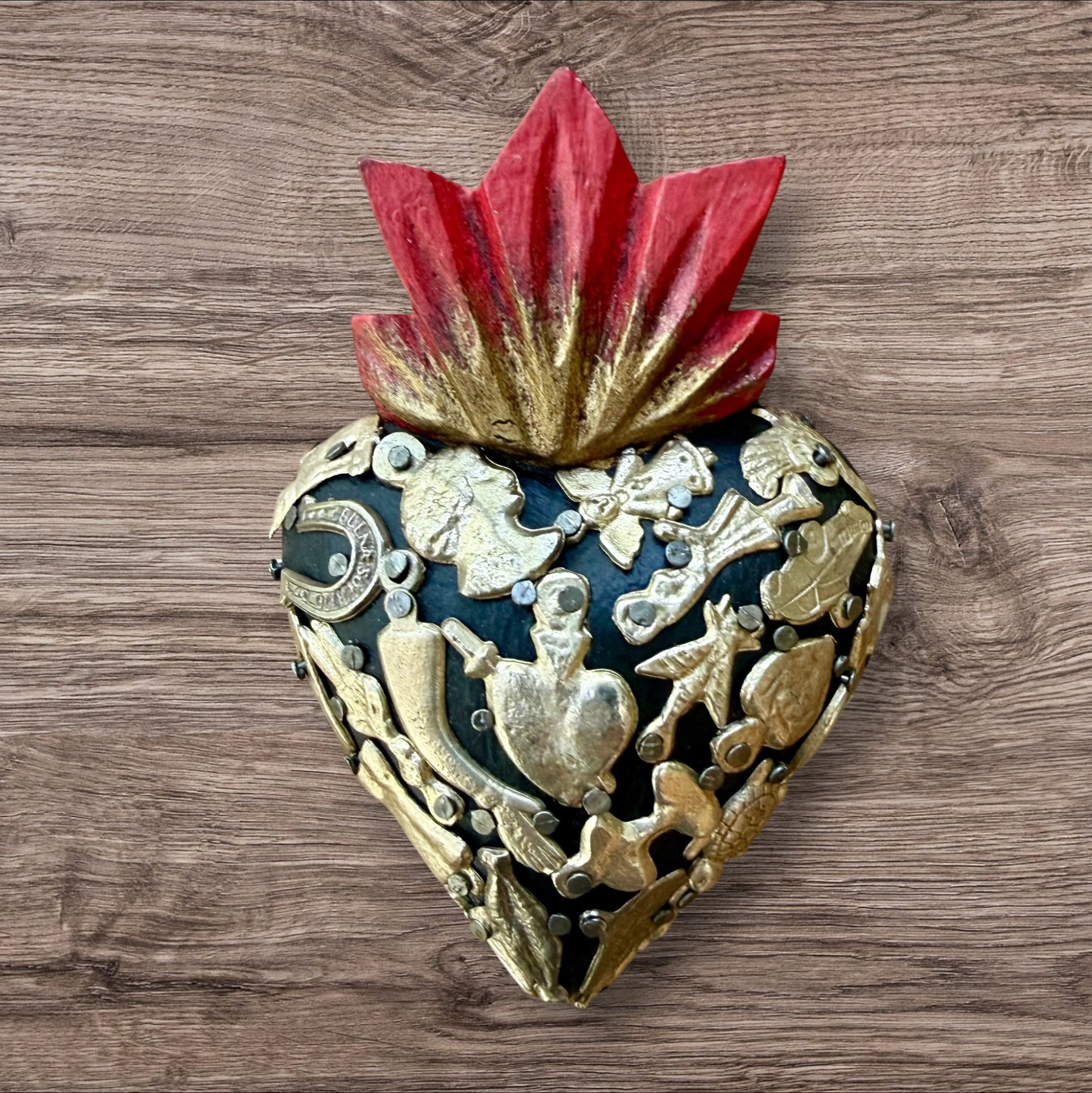A decorative Milagros Sacred Heart featuring a black heart, red flame and antiqued charms, set against a rustic wood background.