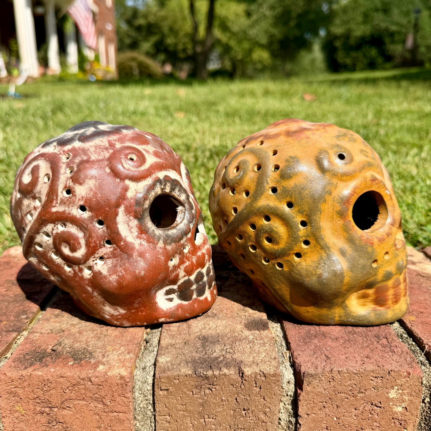 Two decorative clay sugar skull ornaments with Day of the Dead style painted patterns, displayed side-by-side on a brick surface outdoors with a grassy lawn and trees in the background, right side view.