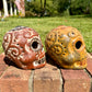 Two decorative clay sugar skull ornaments with Day of the Dead style painted patterns, displayed side-by-side on a brick surface outdoors with a grassy lawn and trees in the background, right side view.