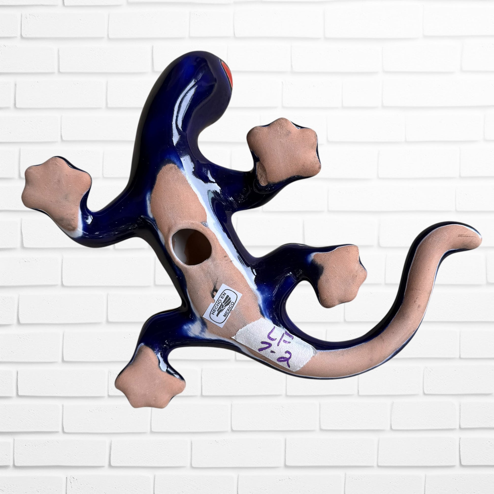 A colorful ceramic wall sculpture of a stylized lizard or salamander, decorated with vibrant hand-painted patterns in blue, red, yellow, and orange, mounted on a white brick wall bottom with hanging hole.