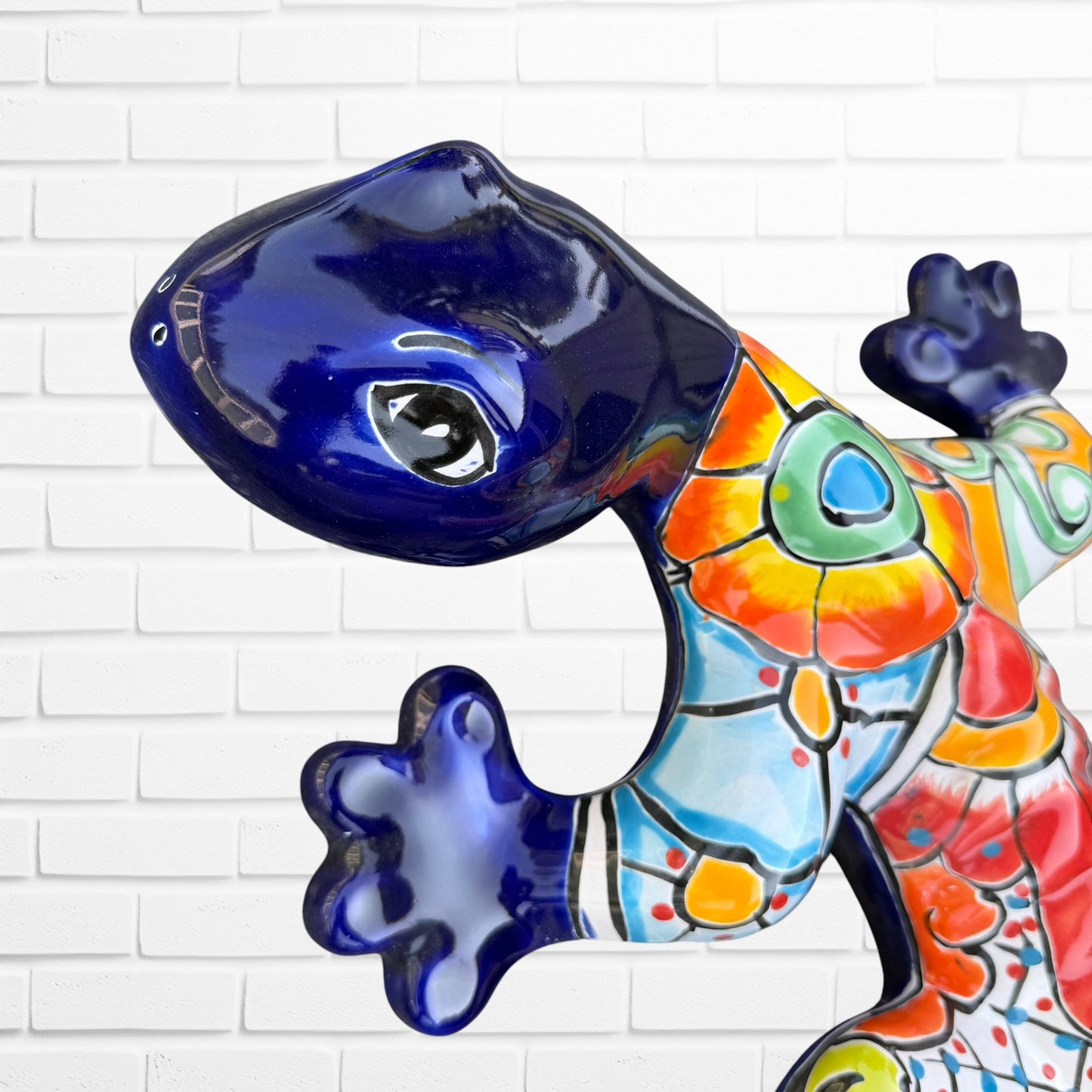 A colorful Talavera ceramic wall sculpture of a salamander, featuring a glossy finish and intricate hand-painted patterns in blue, orange, red, yellow, and white, mounted against a white brick wall,  close-up of the face and eyes.