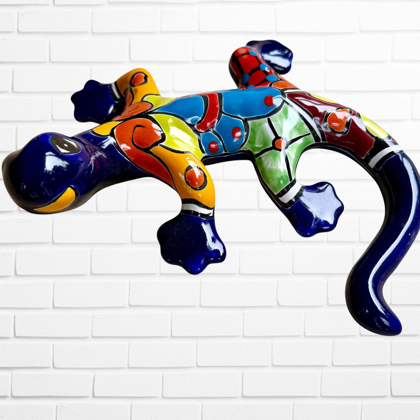 A colorful, glossy lizard wall sculpture with 
a vibrant mosaic pattern of blue, orange, yellow, green, red, and black, mounted on a white brick wall front view
