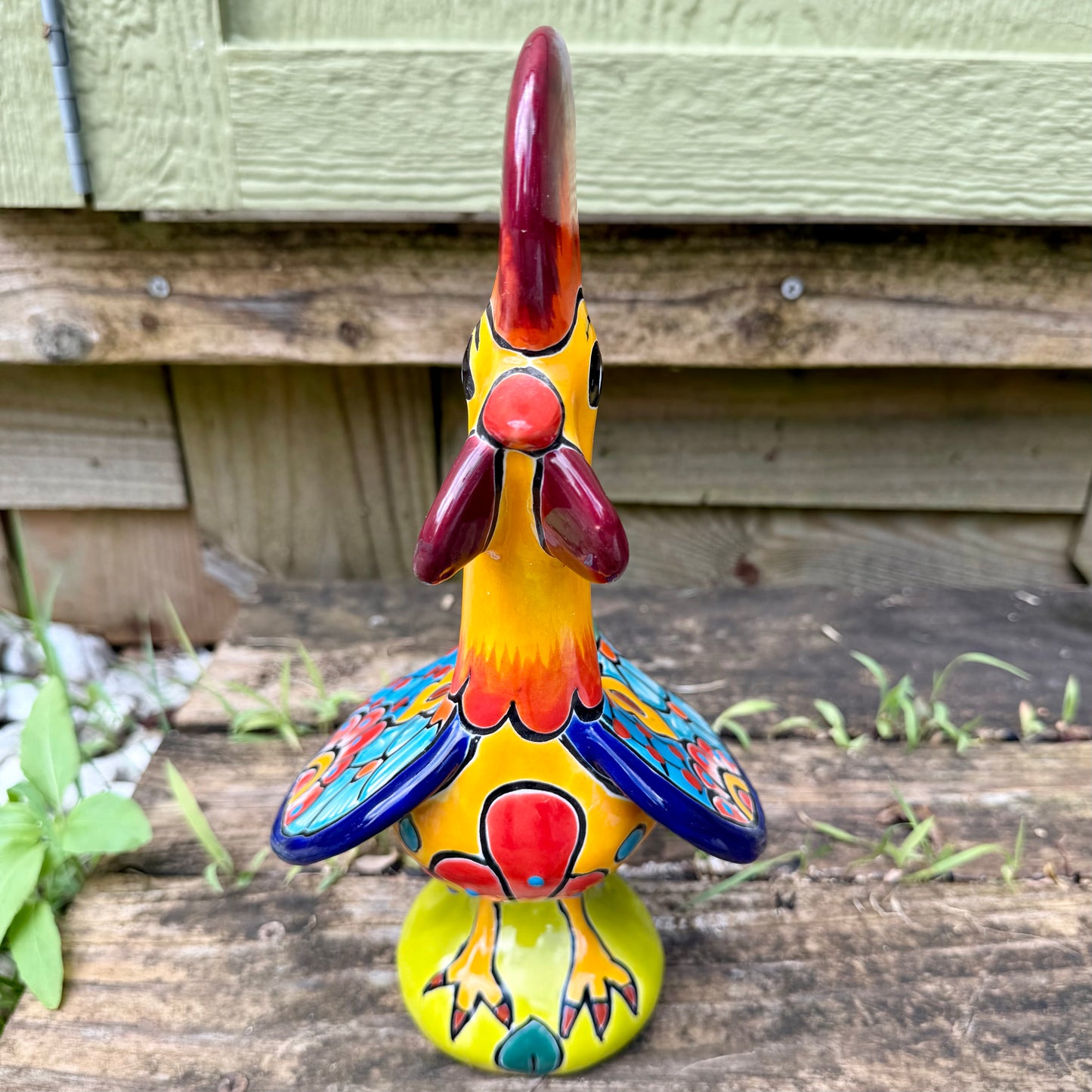 A colorful hand-painted ceramic Mexican-style rooster figurine standing on a rustic wooden surface outdoors, front view.