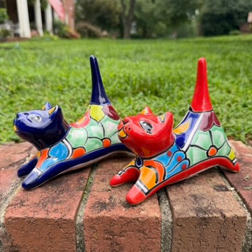 Two colorful ceramic cat figurines, one blue and one red, decorated with geometric patterns, displayed on a brick ledge with a green lawn in the background right side view of the stretch.