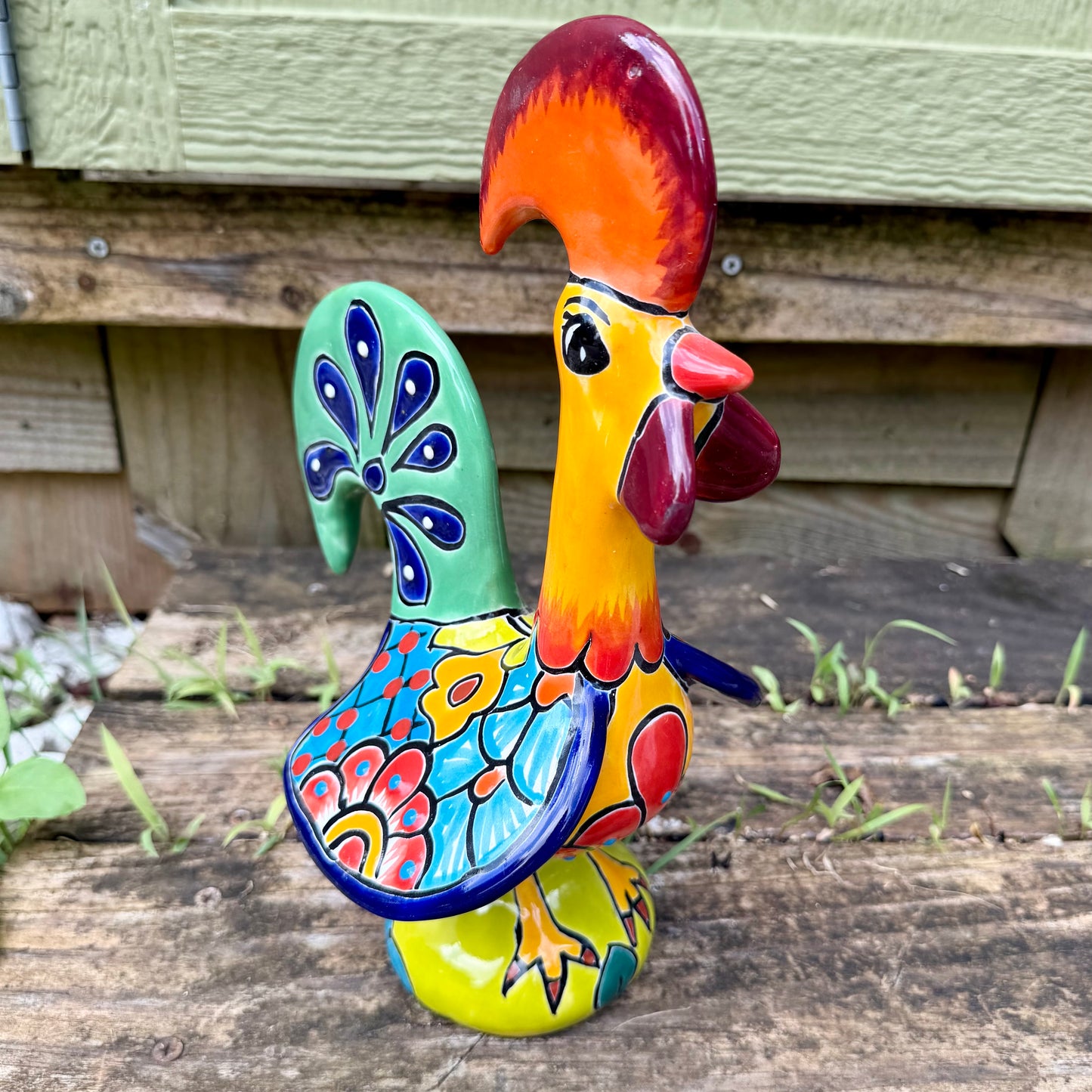 A colorful hand-painted ceramic Mexican-style rooster figurine standing on a rustic wooden surface outdoors.