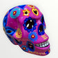 A hand-painted ceramic sugar skull mask in vibrant magenta pink, decorated with colorful floral and geometric patterns in blue, yellow, red, and white, set against a plain white background.