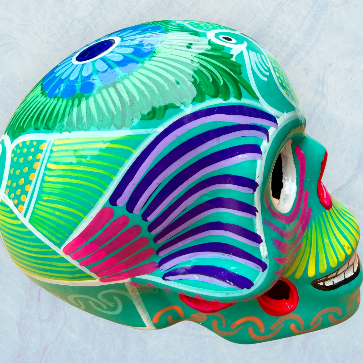 A decorative skull sculpture with intricate colorful sugar skull style patterns painted on a teal colored base, set against a light blue textured background, left side view.