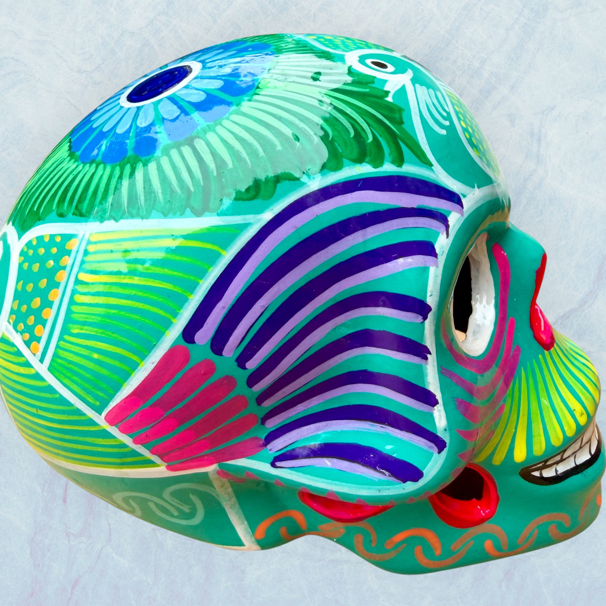A decorative skull sculpture with intricate colorful sugar skull style patterns painted on a teal colored base, set against a light blue textured background, left side view.