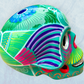 A decorative skull sculpture with intricate colorful sugar skull style patterns painted on a teal colored base, set against a light blue textured background, left side view.