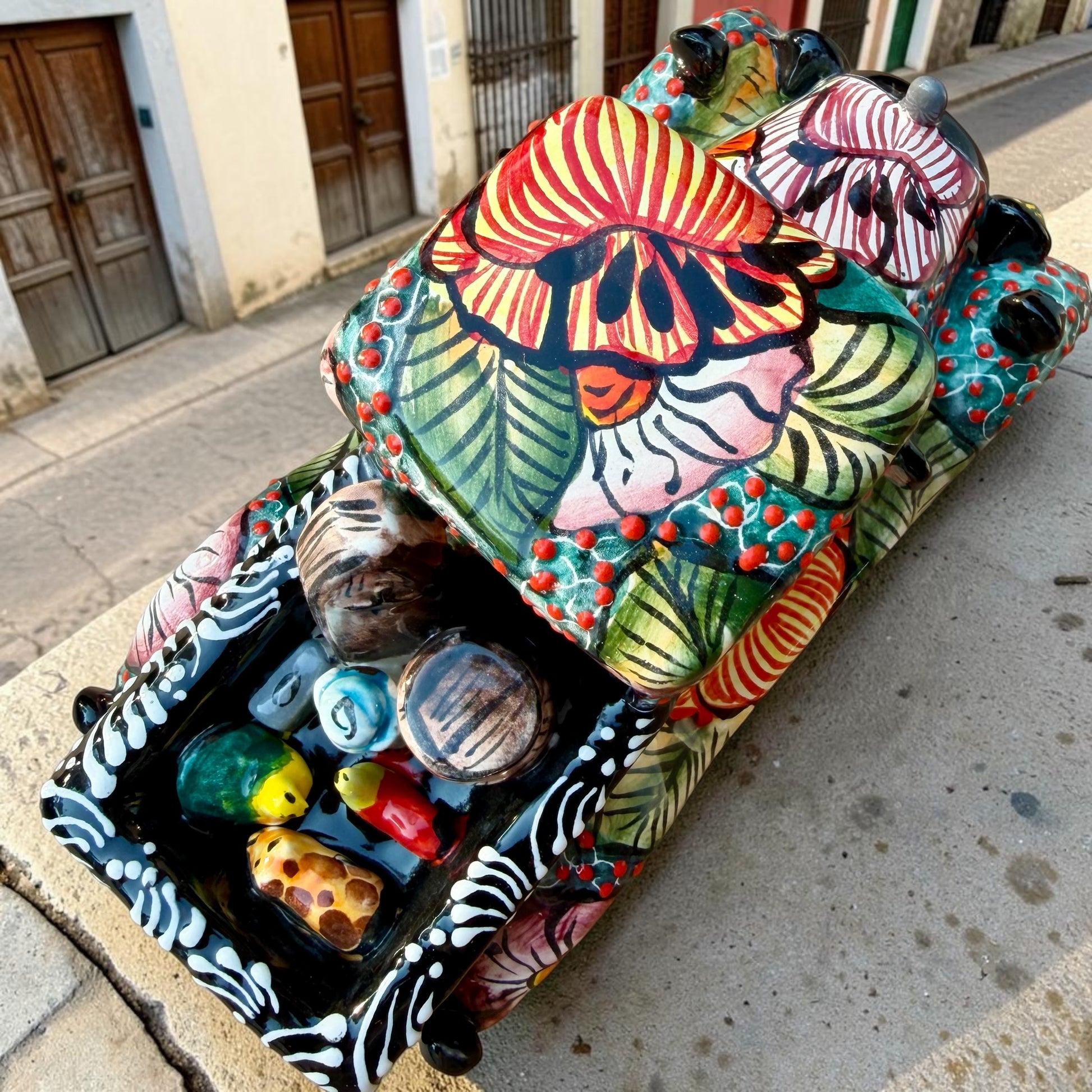 A decorative Talavera day of the dead pick up truck figurine two skeleton passengers. The truck is vibrant, with a hand-painted tropical floral pattern, photographed outdoors on a stone-paved street. This is the top view of the truck showing the bed and animal passengers.