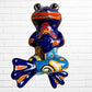 A colorful ceramic frog sculpture sitting cross-legged against a white brick wall background. The frog features a glossy deep blue body with vibrant orange, red, yellow, and light blue painted patterns and details.