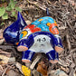 A colorful ceramic cat figurine with a glossy blue head and tail, yellow ears and paws, and a patterned body, standing outdoors on mulch with green plants in the background, bottom on cat.