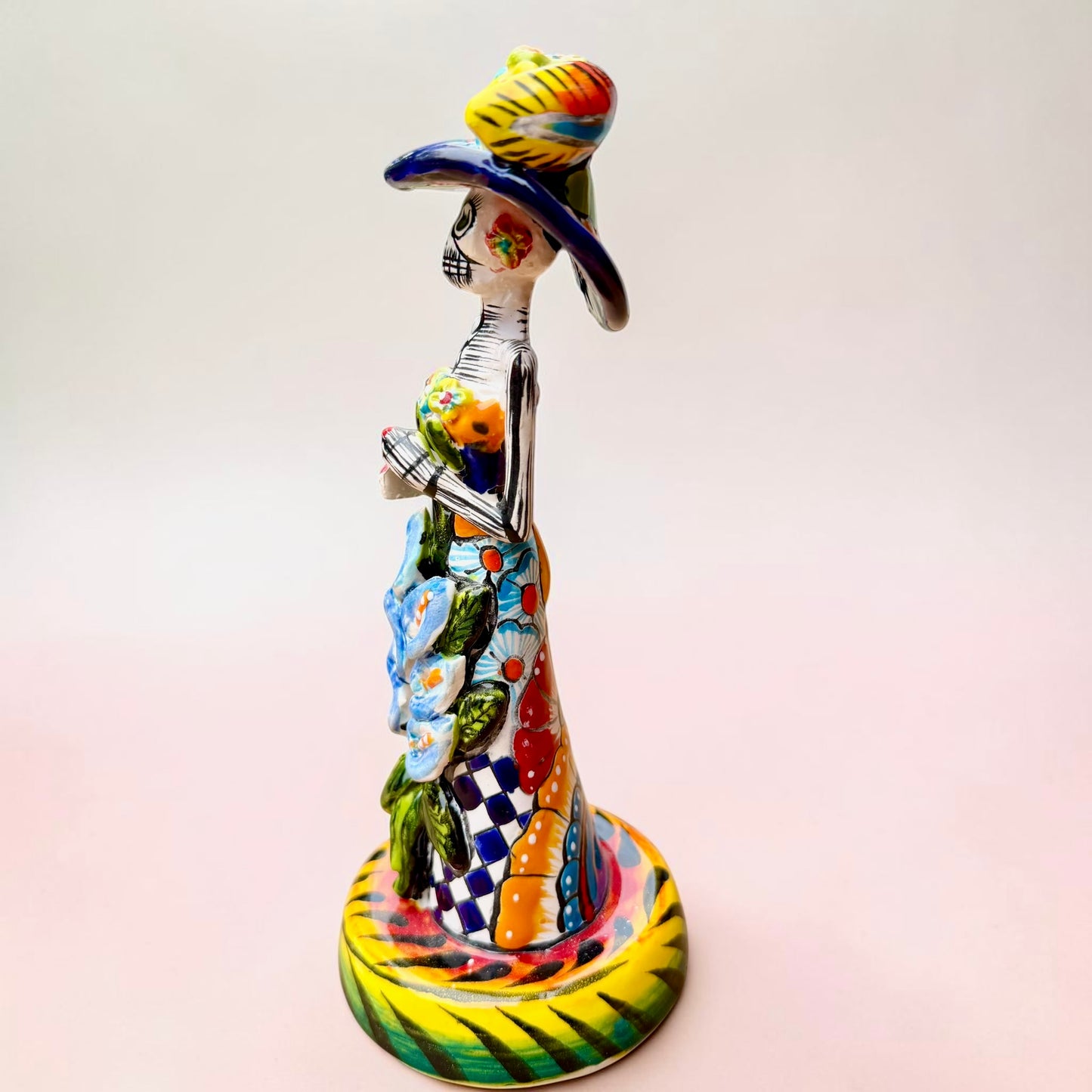 A Talavera Day of the Dead (Día de los Muertos) ceramic skeleton figurine with calla Lillies standing on a patterned base against a soft pink gradient background. Right side view.