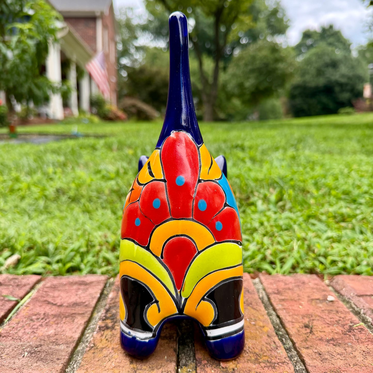 A glossy talavera ceramic cat figurine decorated with vibrant hand-painted patterns in blue, orange, yellow, and red, standing on a brick path with a green lawn and a house in the blurred background. Tail back view.