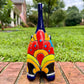 A glossy talavera ceramic cat figurine decorated with vibrant hand-painted patterns in blue, orange, yellow, and red, standing on a brick path with a green lawn and a house in the blurred background. Tail back view.