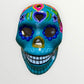 A decorative sugar skull painted in bright turquoise blue with colorful floral and geometric patterns in pink, yellow, green, purple, and white, shown against a plain light background front