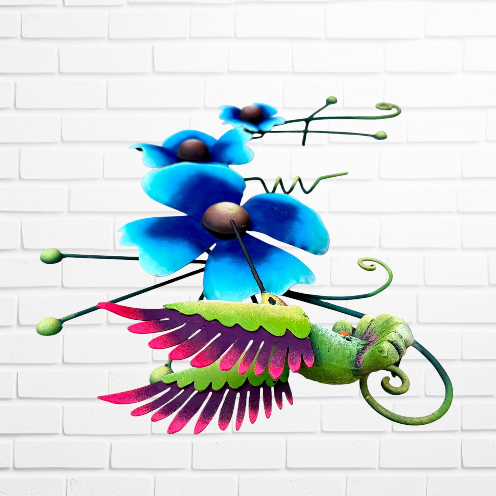 Decorative metal wall art with three blue flowers and a colorful hummingbird on a white brick background bird three dimensional view.