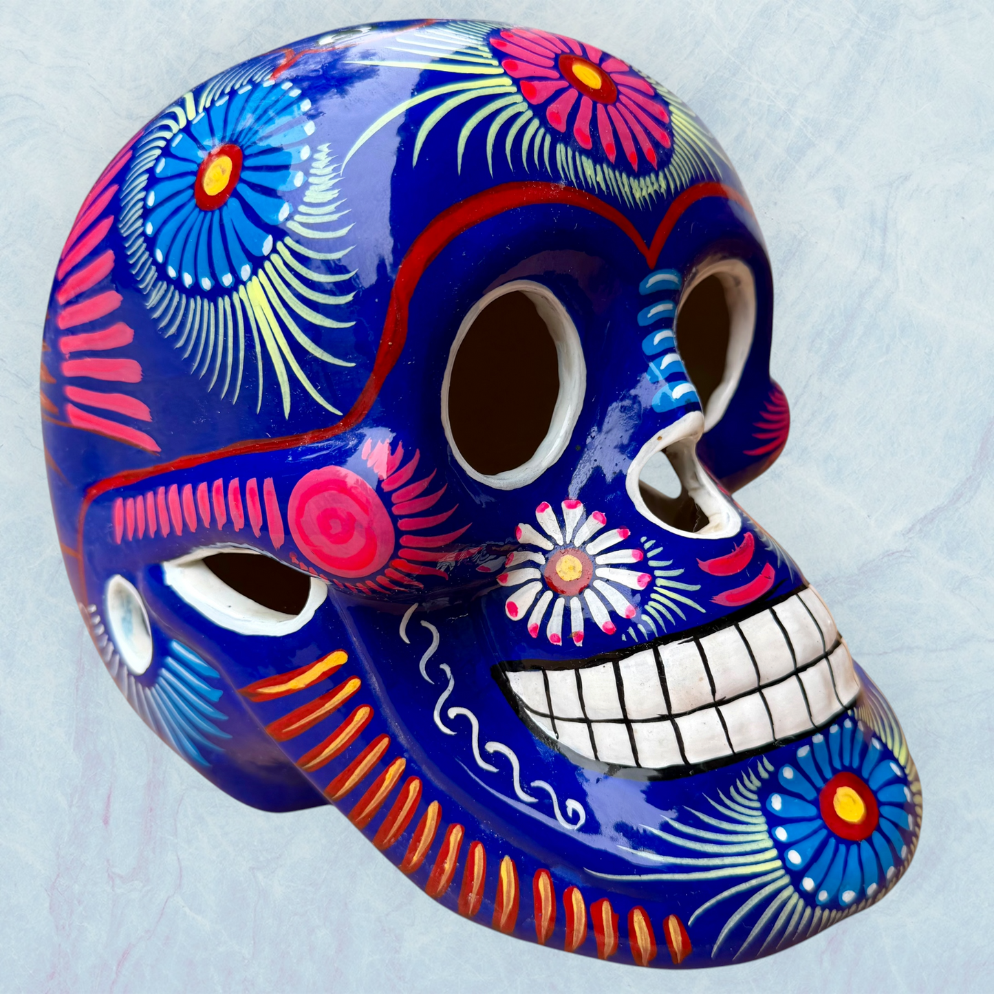 A glossy blue Day of the Dead sugar skull XGrande decorated with vibrant pink, blue, white, and orange floral and geometric patterns, shown against a light blue textured background.