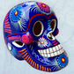 A glossy blue Day of the Dead sugar skull XGrande decorated with vibrant pink, blue, white, and orange floral and geometric patterns, shown against a light blue textured background.