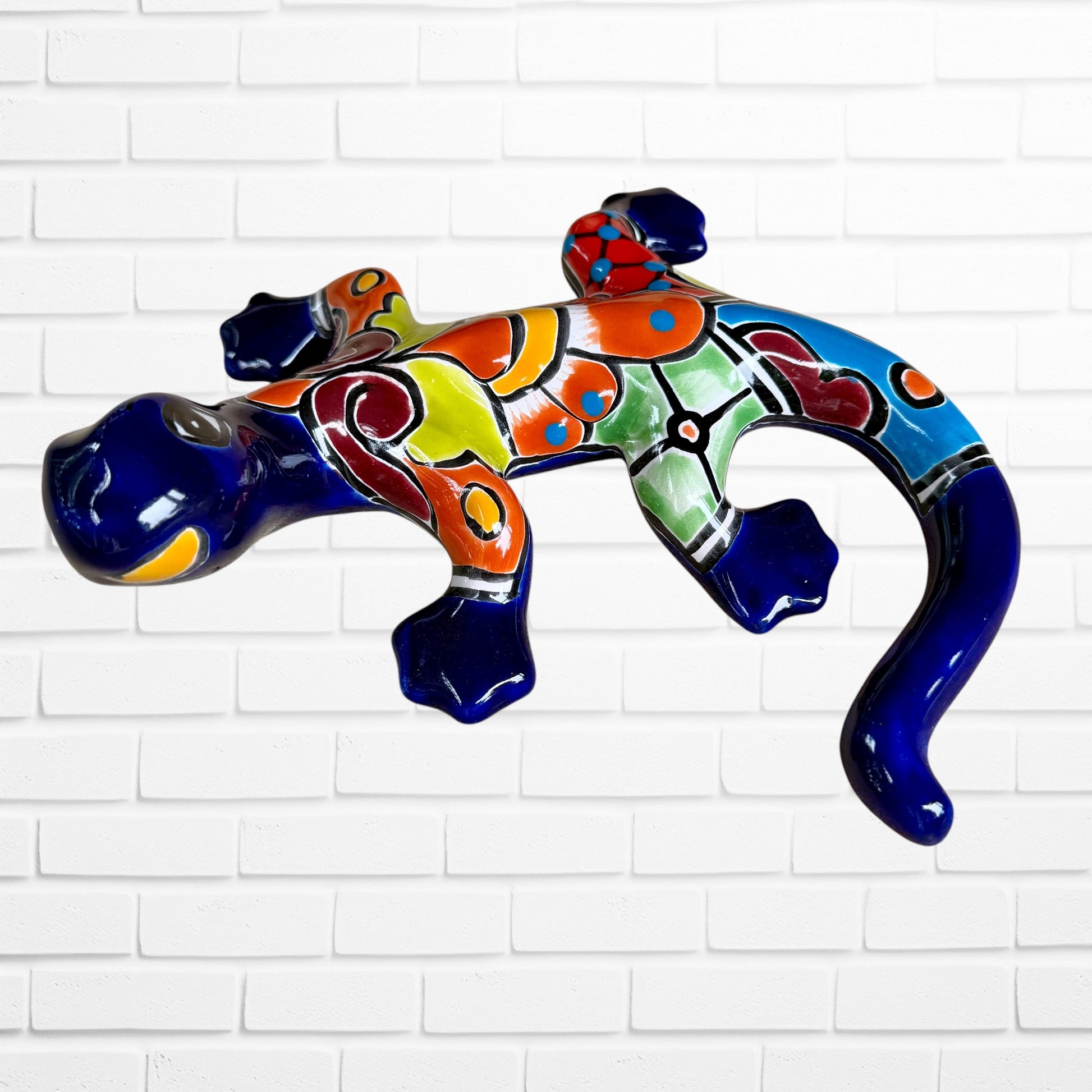 A colorful glazed ceramic lizard wall sculpture with a vibrant multicolor pattern, displayed against a white brick wall background front face view.