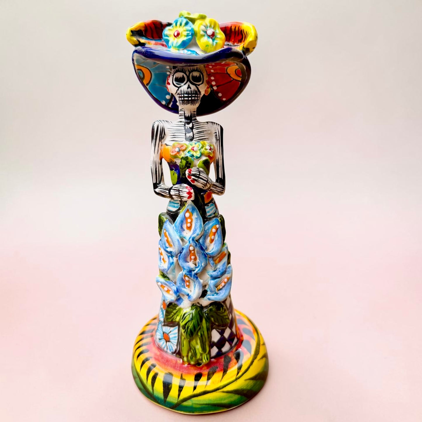 A Talavera Day of the Dead (Día de los Muertos) ceramic skeleton figurine with calla Lillies standing on a patterned base against a soft pink gradient background.