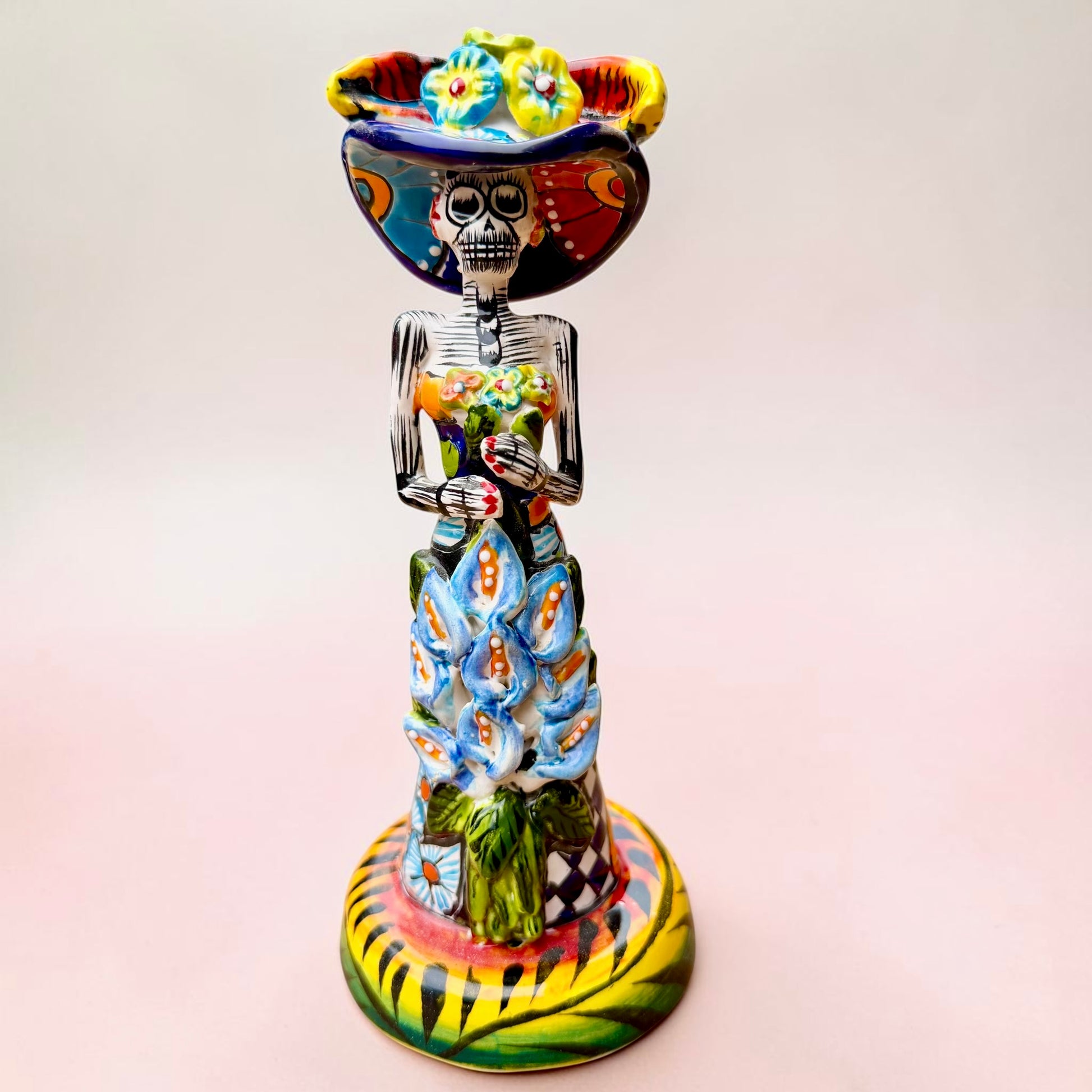 A Talavera Day of the Dead (Día de los Muertos) ceramic skeleton figurine with calla Lillies standing on a patterned base against a soft pink gradient background.