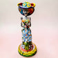 A Talavera Day of the Dead (Día de los Muertos) ceramic skeleton figurine with calla Lillies standing on a patterned base against a soft pink gradient background.