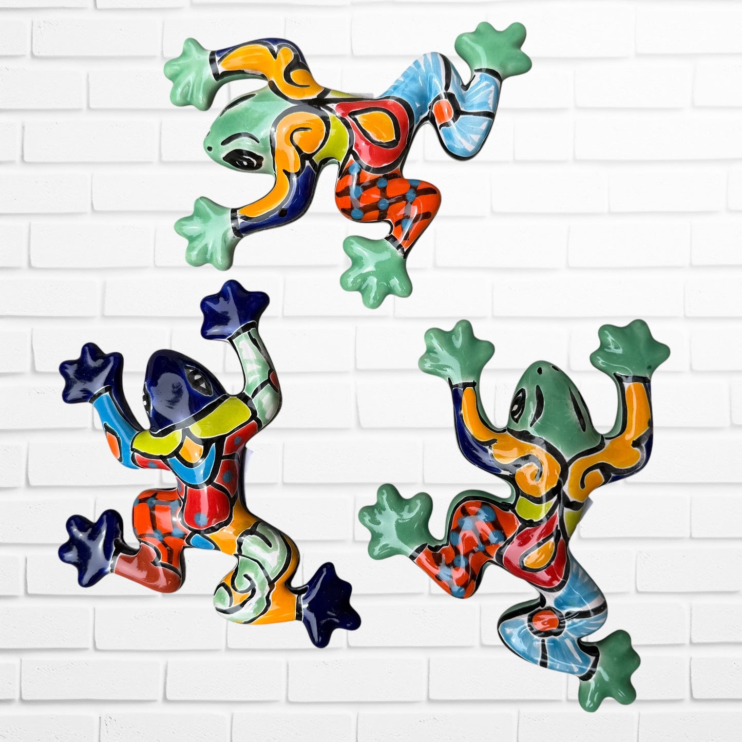 Three colorful ceramic wall-mounted frog sculptures in a vibrant multicolor design, displayed against a white brick wall background.