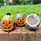 Three hand-painted Talavera ceramic jack-o'-lantern pumpkins with orange faces and yellow, blue, and red green designs, sitting on a brick path in a grassy backyard, bottom view.