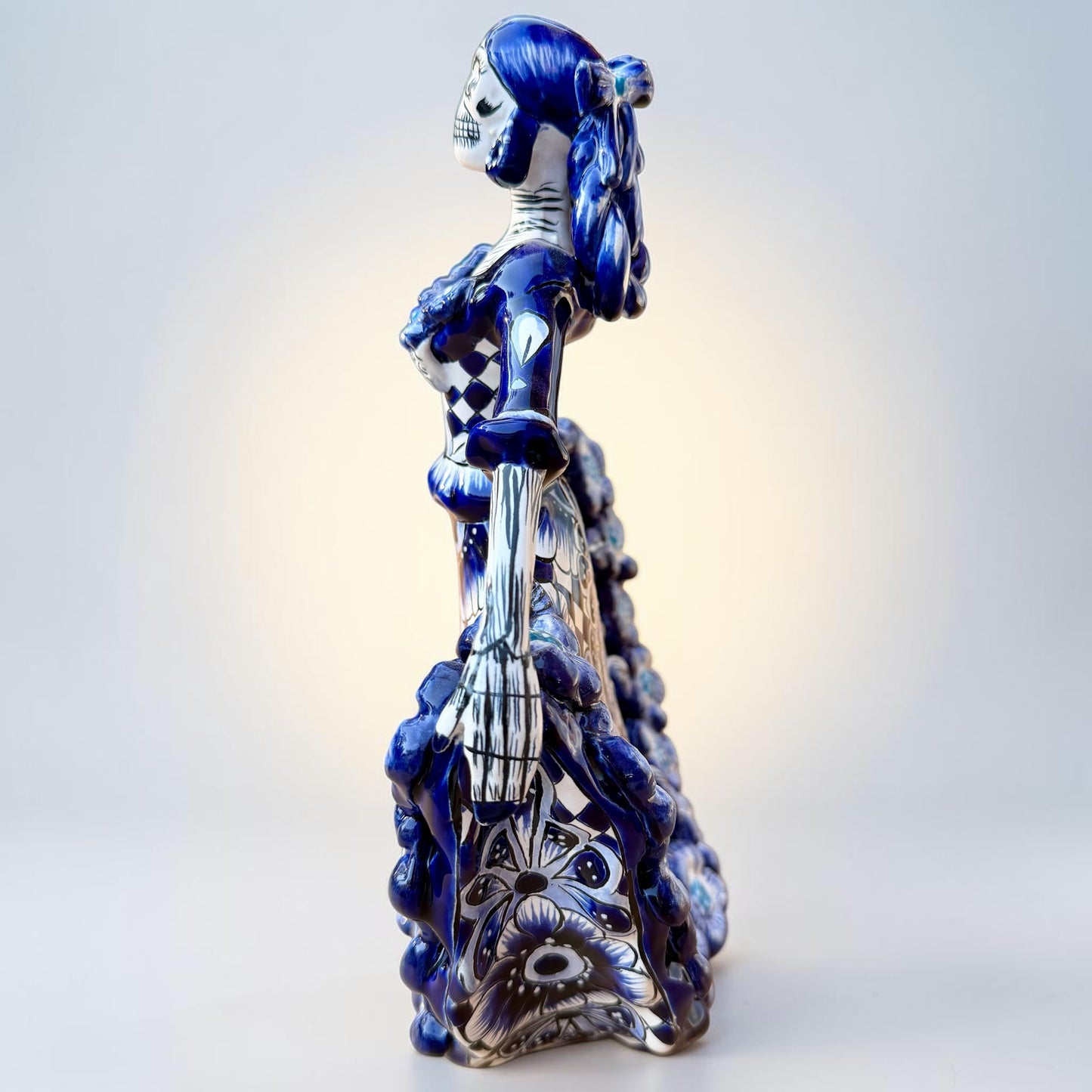 A Day of the Dead style talavera figurine of a skeleton woman in a wide floral skirt, painted in deep blue, black, white, and light blue on a soft beige-to-blue gradient background. Right side view of ballerina.