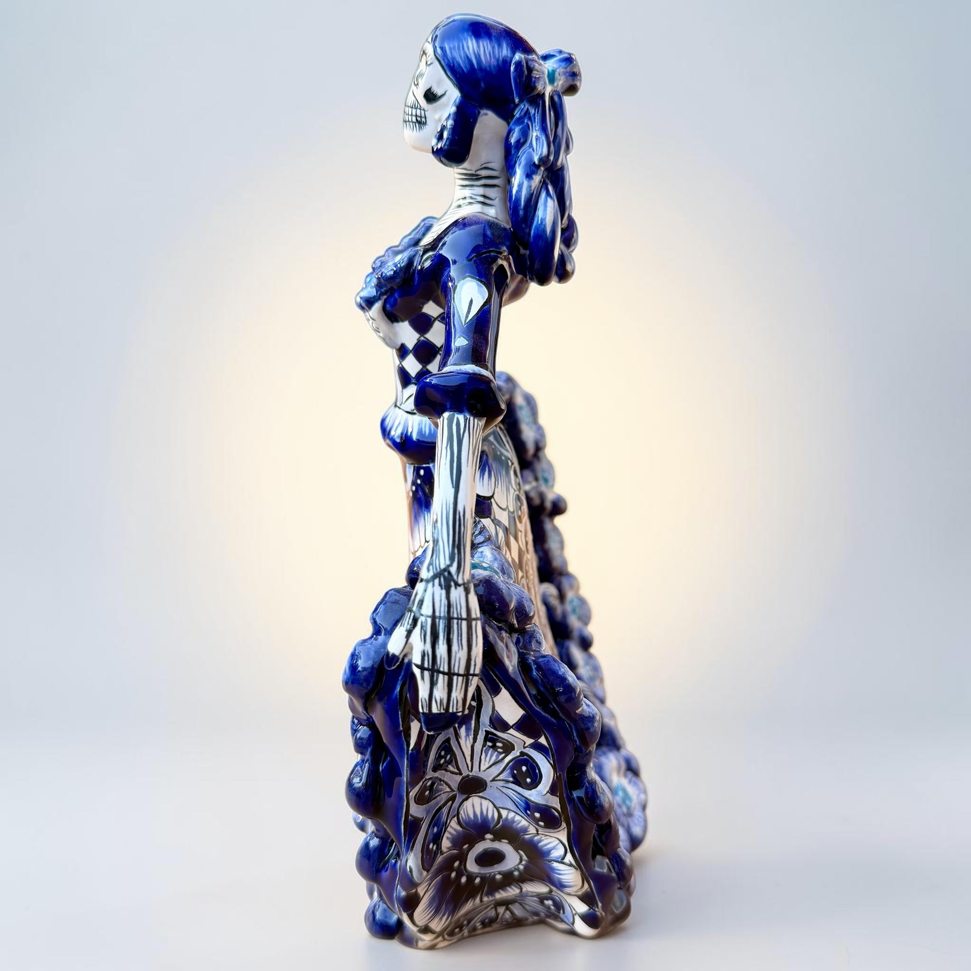 A Day of the Dead style talavera figurine of a skeleton woman in a wide floral skirt, painted in deep blue, black, white, and light blue on a soft beige-to-blue gradient background. Right side view of ballerina.