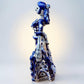 A Day of the Dead style talavera figurine of a skeleton woman in a wide floral skirt, painted in deep blue, black, white, and light blue on a soft beige-to-blue gradient background. Right side view of ballerina.