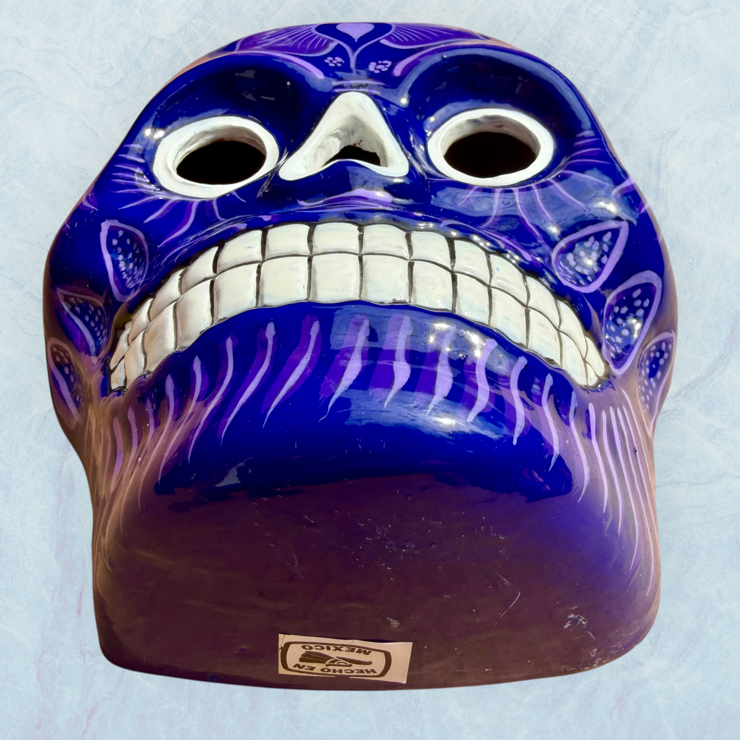 A clay, purple with designs, Mexican-style skeleton skull face mask hanging on a soft blue surface, view from the chin.