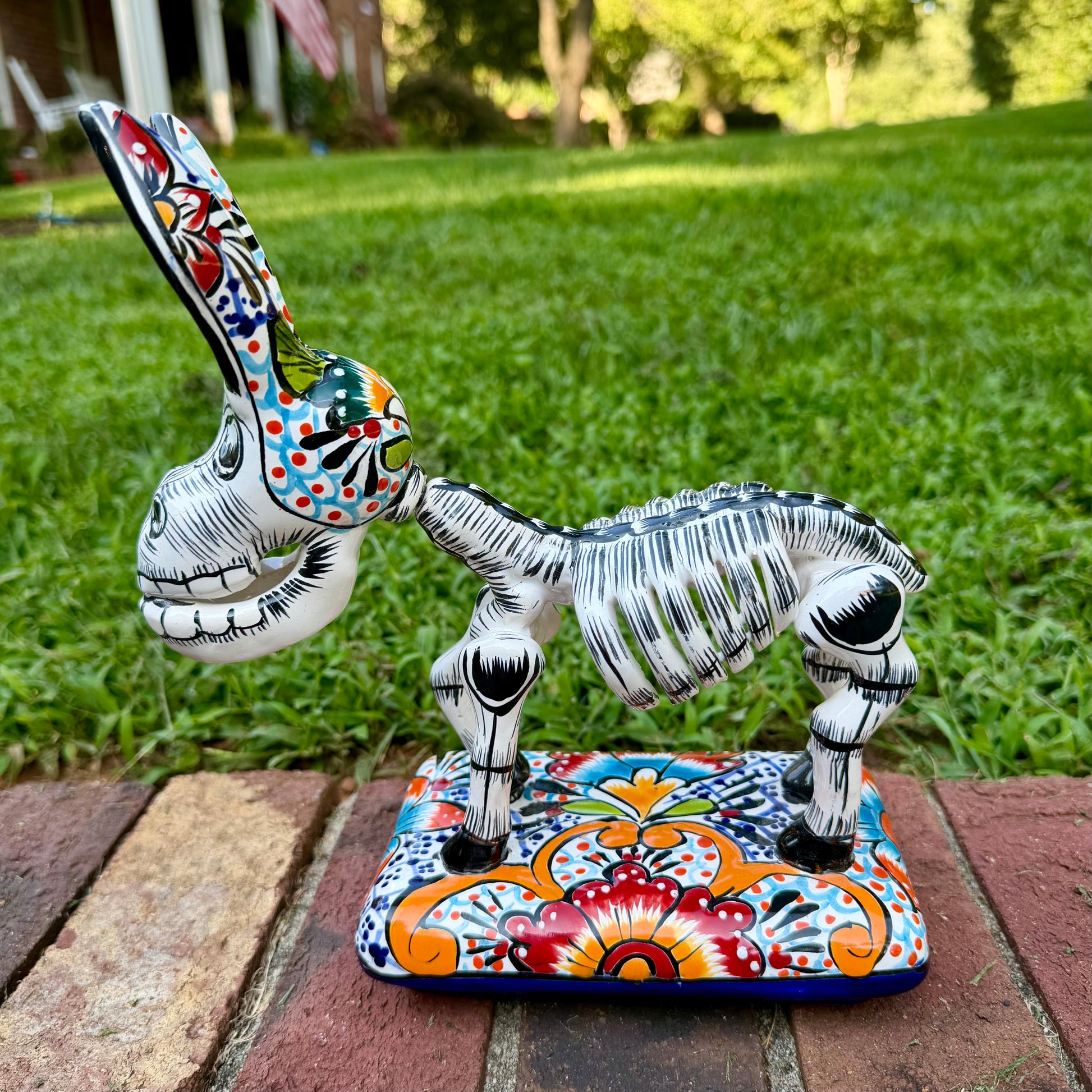 A hand-painted Mexican Talavera-style donkey figurine with a skeletal design, standing on a colorful floral base outdoors on a brick path with a green lawn in the background. This is the right side view of the donkey.
